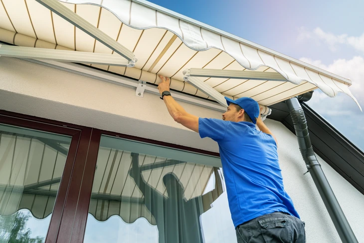 Technicien en tenue bleue installant un store banne rétractable sur la terrasse d'une maison résidentielle