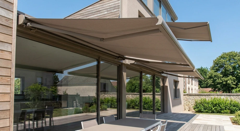 Terrasse résiduelle spacieuse avec stores enroulables de couleur ivoire ou taupe, déployés en position fermée au-dessus de baies vitrées, installés sur la façade d'une maison contemporaine. Vue d'angle montrant clairement la pose professionnelle du système sur le bâtiment. La terrasse adjacent est partiellement visible avec mobilier extérieur moderne. Photographie en lumière naturelle de jour, mettant en évidence la finition de qualité et l'effet d'occultation créé par les stores.