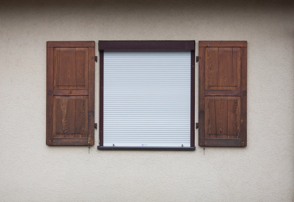 Façade extérieure de bâtiment avec fenêtre équipée d'un volet roulant blanc fermé, encadrée de deux volets battants en bois massif de teinte brune