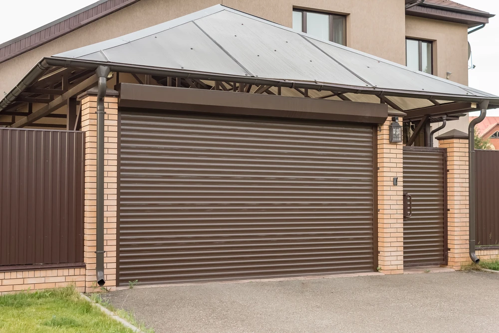 Garage résidentiel extérieur avec porte sectionnelle coulissante en lamelles métalliques brunes, structure en briques beiges sous charpente apparente et toiture métallique grise