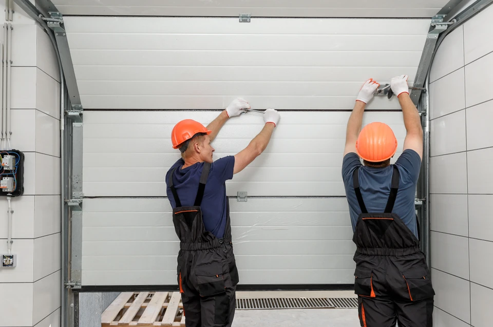 Deux techniciens en combinaisons de travail et casques orange installent une porte de garage sectionnelle blanche dans un garage résidentiel