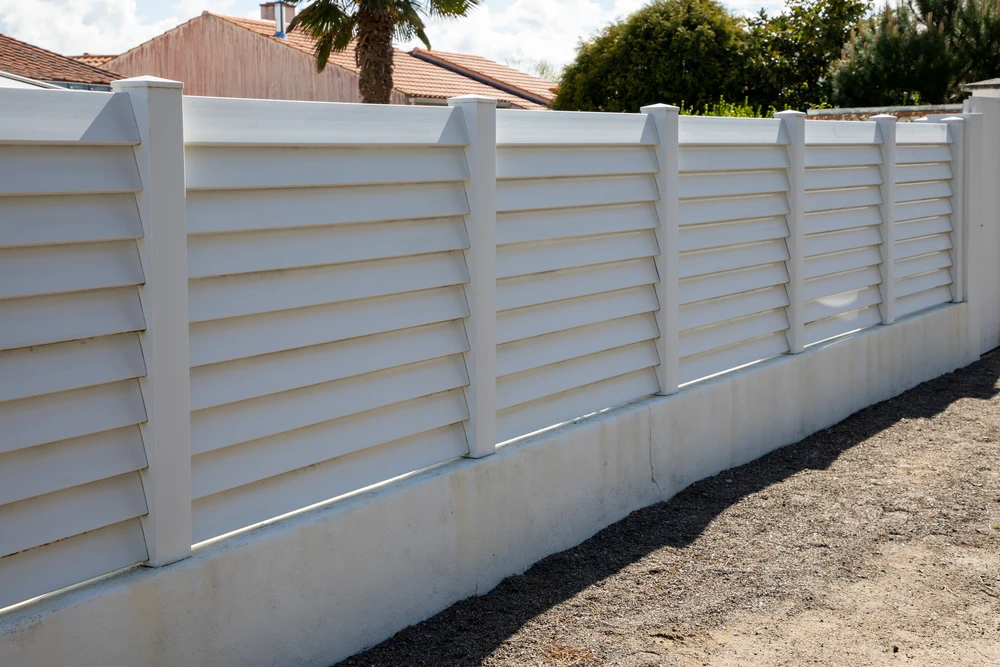 Clôture extérieure résidentielle avec panneaux à lames horizontales blanches orientables montés sur poteaux verticaux, installée sur muret béton bas