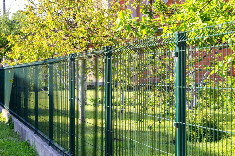 Clôture grillagée rigide verte installée le long d'une propriété résidentielle, délimitant un terrain arboré avec végétation dense et maison en arrière-plan