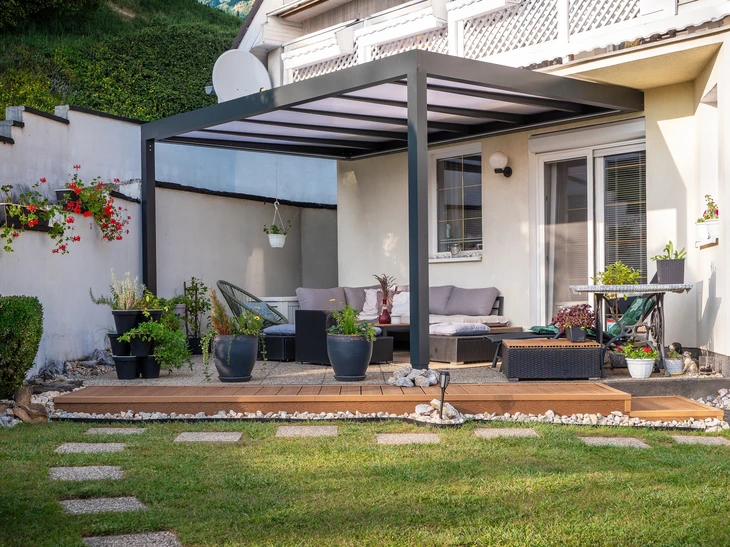 Terrasse couverte avec pergola en bois et colonnes blanches, mobilier de jardin en résine tressée avec coussins gris, donnant sur pelouse et aire de jeux