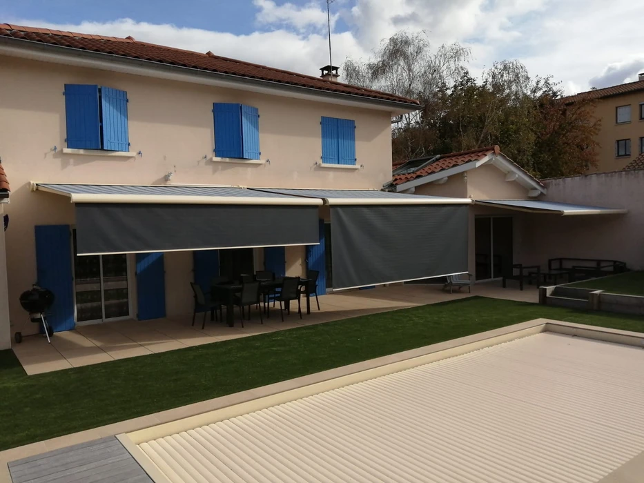 Volet roulant ou store enroulable beige/gris clair installé sur la fenêtre d'une véranda moderne, partiellement déployé pour créer une zone ombragée à l'intérieur. La véranda possède une structure en aluminium et des vitrages, avec vue sur un jardin en arrière-plan. La photo montrant clairement la pose professionnelle du système de fermeture intégré à la structure de la véranda, photographiée en lumière naturelle de jour pour mettre en évidence la qualité de l'installation et l'effet d'occultation créé.